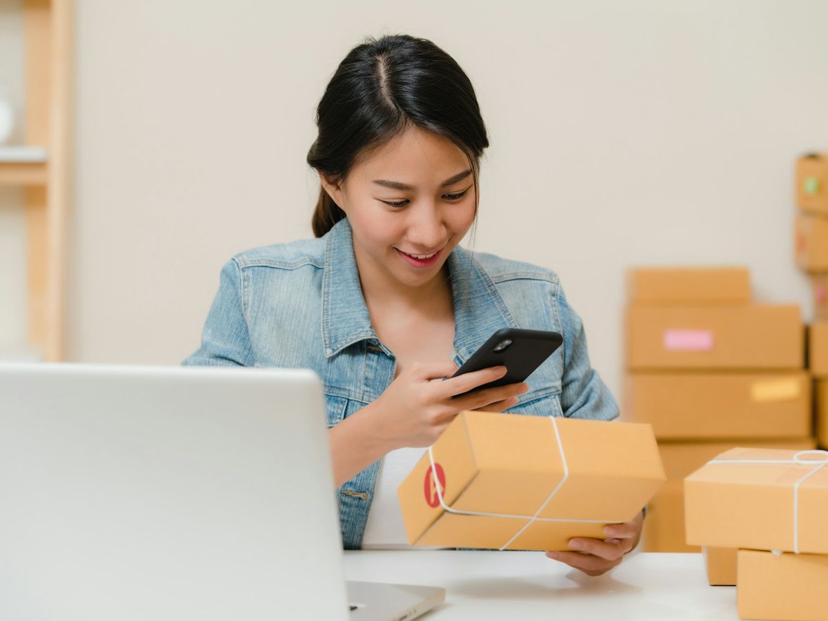 Empreendedora conferindo pedidos no celular, ao lado de encomendas embaladas para envio.